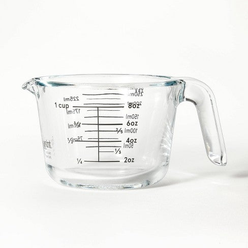 A clear glass measuring cup with a handle and spout, showing measurement markings in cups, ounces, and milliliters on the side, placed against a plain white background.