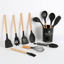 A set of black kitchen utensils with wooden handles, including spatulas, a whisk, tongs, a pasta server, a basting brush, a spoon, and a ladle, displayed beside and inside a black container.