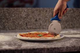 A hand holding a pizza cutter slices a thin-crust pizza with tomato sauce and basil on a white plate, placed on a floured granite countertop.