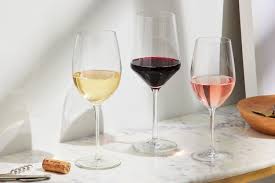 Three wine glasses on a marble surface, filled with white wine, red wine, and rosé. A corkscrew and cork are nearby, with a wooden bowl partially visible on the right. Bright, natural lighting.
